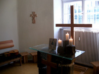Bild vom Andachtsraum, im Vordergrund sieht man einen Altar aus Steinen mit Glasplatte und drei brennenden Kerzen darauf und einer Ikone. Im Hintergrund sieht man ein großes Holzkreuz