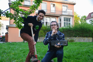 Zu sehen sind zwei Menschen im Garten, einer steht auf einem Spaten, der andere hält eine Kasse in der Hand und deutet mit dem Zeigefinger vor den Lippen an leise zu sein. 